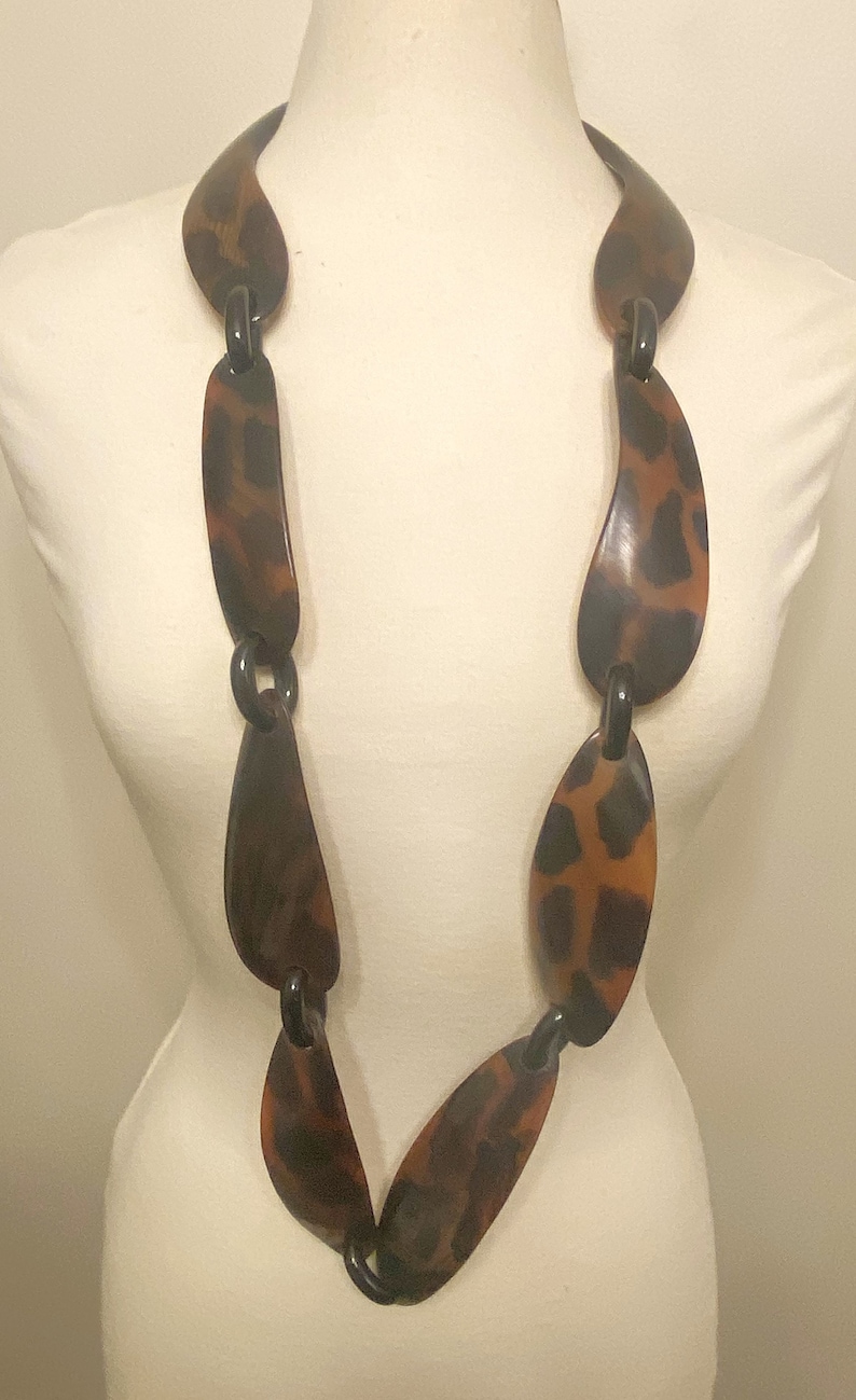 Peut inclure: Un long collier brun et noir compos&eacute; de grandes pi&egrave;ces ovales. Les pi&egrave;ces sont reli&eacute;es par des anneaux m&eacute;talliques noirs.