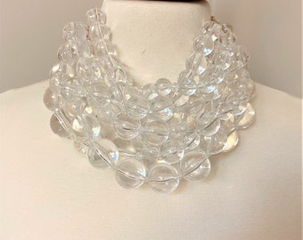 Peut inclure: Un collier imposant à plusieurs rangs composé de nombreuses perles sphériques transparentes de tailles variées. Les perles, semblables à du verre, sont disposées en plusieurs couches, créant un aspect volumineux et groupé.