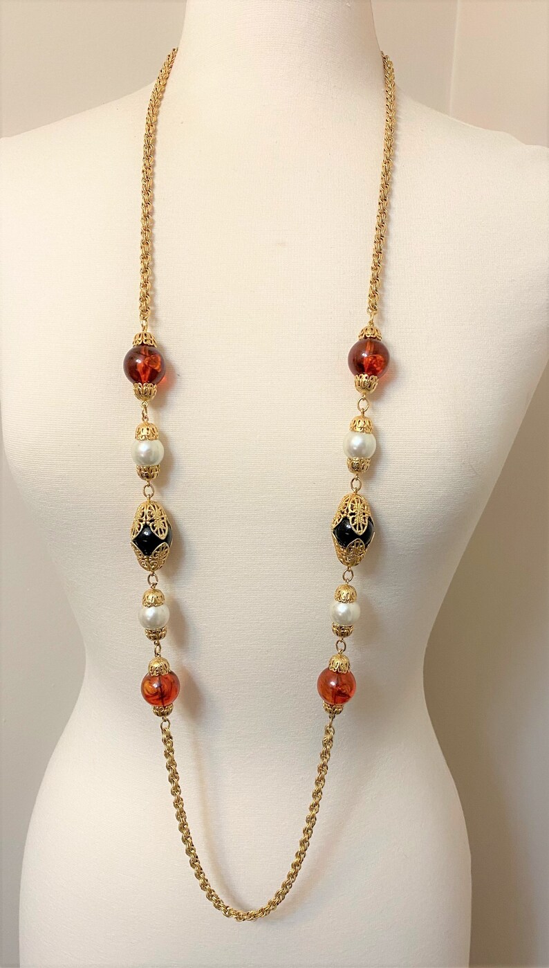 May include: A long gold chain necklace with alternating amber, white pearl, and black and gold beads.
