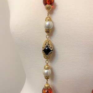 May include: A long gold chain necklace with alternating amber, black, and white beads. The beads are all different shapes and sizes, and the chain is made of small gold links.