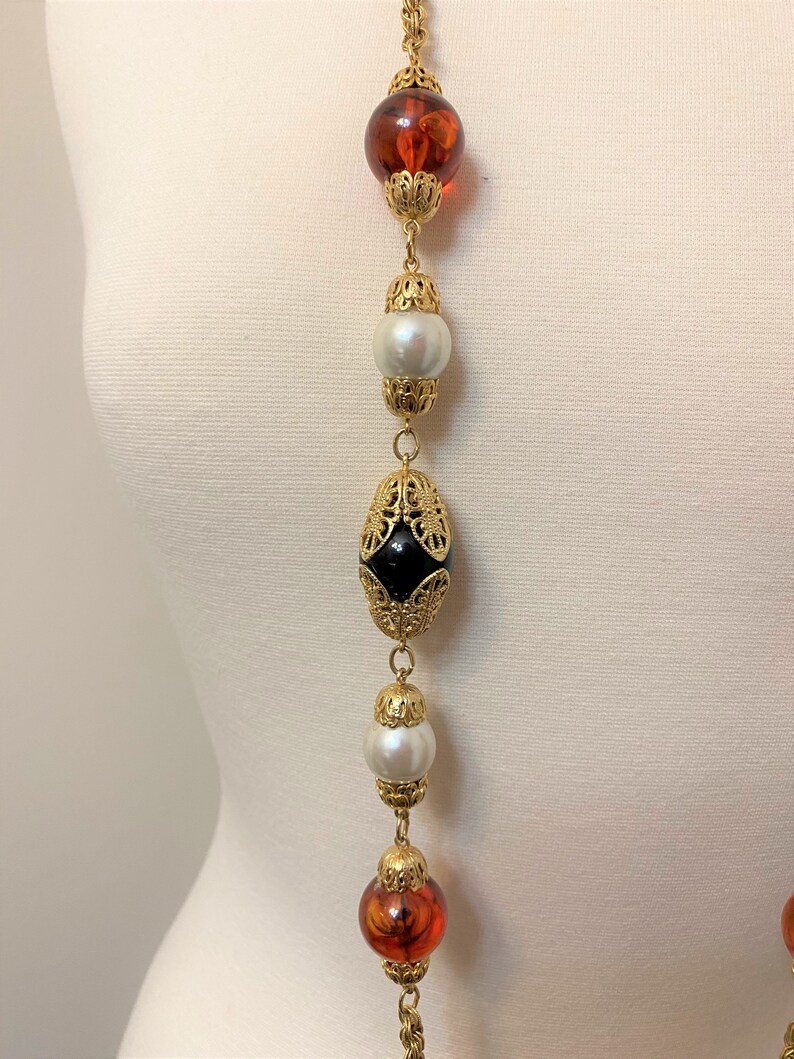 May include: A long gold chain necklace with alternating amber, black, and white beads. The beads are all different shapes and sizes, and the chain is made of small gold links.