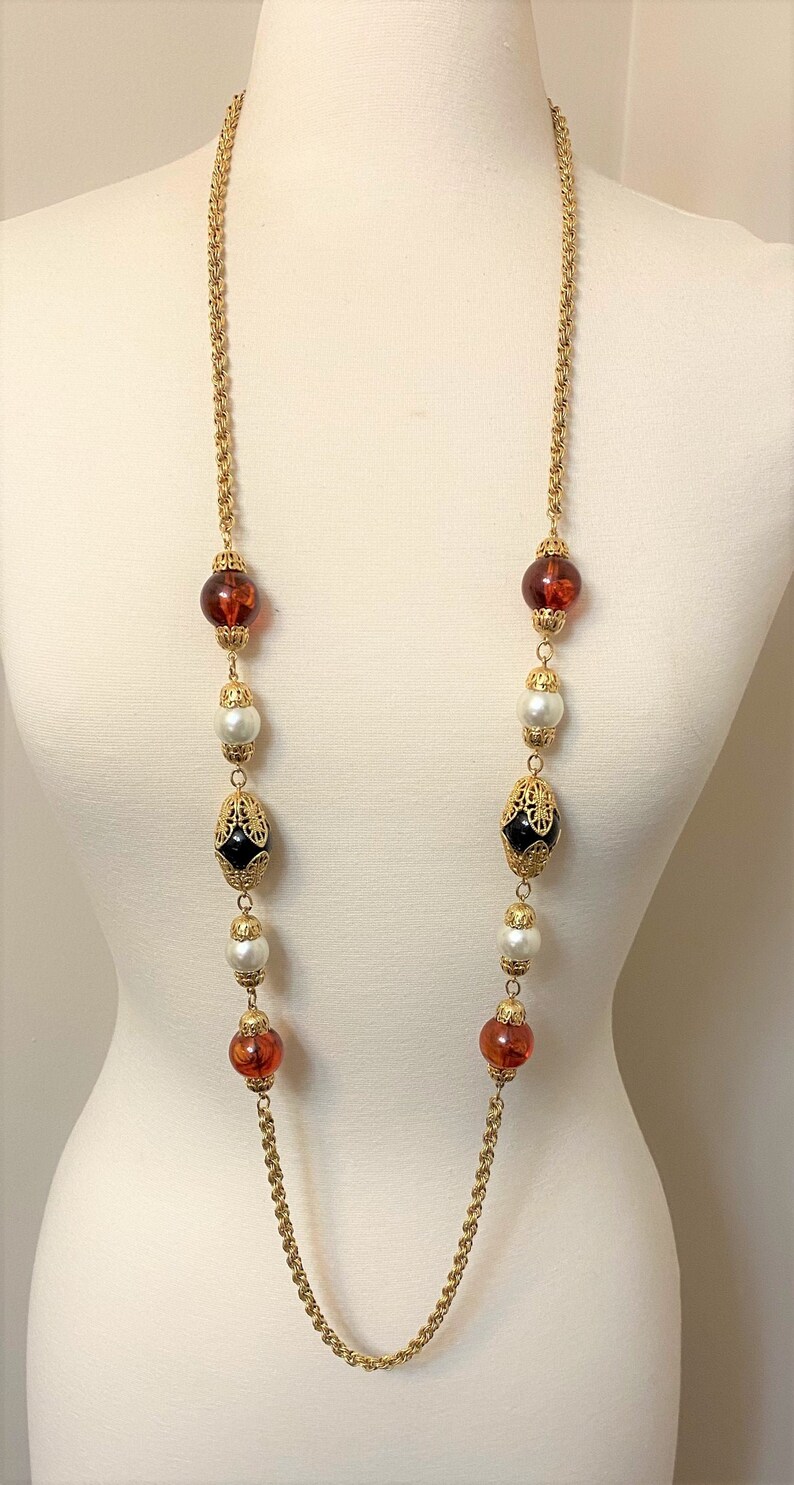 May include: A long gold chain necklace with alternating amber, white pearl, and black and gold filigree beads.