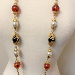 May include: A long gold chain necklace with alternating black and amber glass beads, white faux pearls, and gold filigree accents.