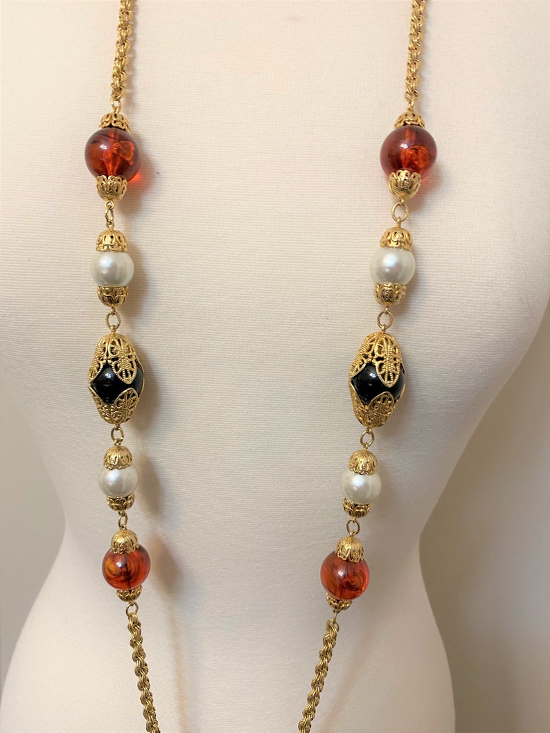 May include: A long gold chain necklace with alternating black and amber glass beads, white faux pearls, and gold filigree accents.