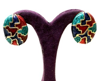May include: Oval-shaped earrings with a colorful, abstract design. The earrings feature a mosaic pattern in red, teal, navy, and gold. The earrings are displayed on a purple velvet earring stand against a white background.
