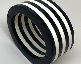 May include: A bold, geometric bangle bracelet featuring alternating black and white stripes. The bracelet has a rounded rectangular shape and a glossy finish, creating a striking contrast. This accessory is a statement piece, perfect for adding a touch of modern flair.