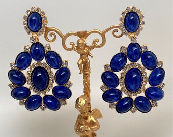May include: A pair of gold tone earrings with blue oval-shaped stones and clear rhinestones. The earrings are hanging from a gold stand.