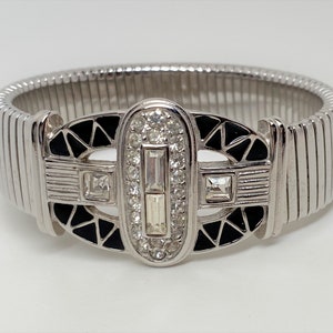 Puede incluir: Un brazalete extensible de plata con un diseño de esmalte negro y pedrería. El diseño presenta un patrón geométrico con una forma ovalada central.