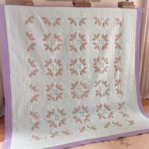 May include: A pale blue quilt featuring a floral design in pink and green. The quilt has a lavender border and a quilted pattern. The floral pattern is arranged in a repeating circular design.