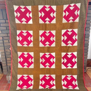 May include: An antique quilt featuring a repeating pattern of red and white geometric shapes on a brown backdrop. The quilt has a green border and a red binding. The quilt is displayed on a wall.