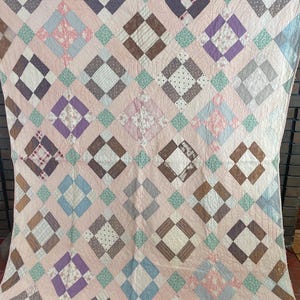 May include: A vintage quilt with a pink background and a diamond pattern. The diamonds are composed of various fabrics in shades of brown, purple, blue, and white. The quilt has a patchwork design and a soft, textured appearance.