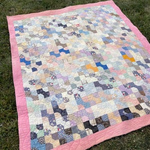 May include: A handmade quilt with a patchwork design in various pastel colors, including blue, pink, and lavender. The quilt features a pink border and is laid out on a grassy surface. The quilt's pattern is a mix of geometric shapes.