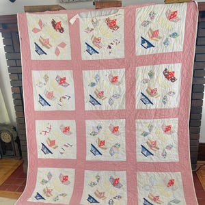 May include: A vintage quilt with a pink border and nine white squares. Each square features a floral design with red, blue, and yellow accents. The quilt is draped over a fireplace.