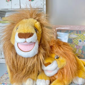 May include: Two plush lion toys. The larger lion has a yellow face, brown nose, and a wide, smiling mouth with a pink tongue. The smaller lion is nestled beside it, with a similar design. Both have shaggy, brown manes.