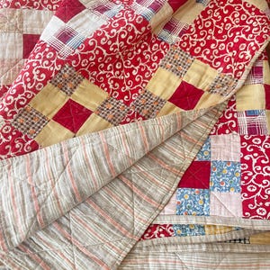 Peut inclure: Une courtepointe faite à la main avec un motif patchwork. La courtepointe présente des carrés de tissus rouges, crème et jaunes. Le tissu rouge a un motif tourbillonnant blanc. Le dessous est un tissu rayé beige et rose.