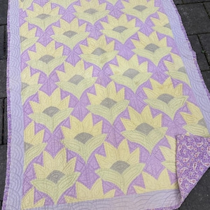 May include: A handmade quilt with a repeating floral design in yellow, grey, and lavender. The quilt has a lavender border and a paisley patterned backing. The quilt is likely made of cotton and is suitable for a bed or as a decorative throw.