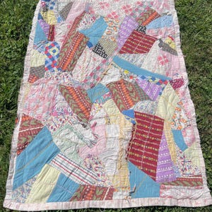 May include: A patchwork quilt with a variety of fabric patterns and colors, including blue, red, pink, and yellow. The quilt has a white border and is laid out on green grass. The quilt is a mix of geometric shapes and patterns.