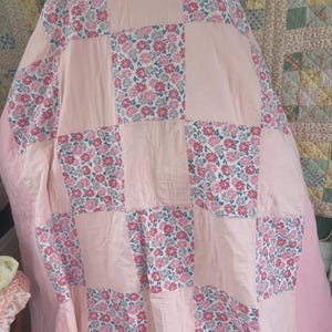 May include: A handmade quilt with a patchwork design. The quilt features alternating squares of solid light pink fabric and floral patterned fabric with pink, white, and grey flowers. A soft, cosy home decor item.