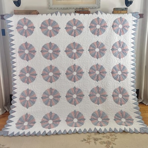 May include: A large, white quilt with a repeating pattern of stylized floral designs in shades of pink and gray. The quilt has a scalloped edge and is displayed on a wooden rod.