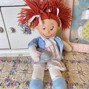 May include: A vintage rag doll with bright red yarn hair styled in pigtails, secured with white ribbons. The doll wears a blue jacket over a white dress with a pink floral pattern and holds a grey stuffed animal. The doll has blue shoes and white socks.
