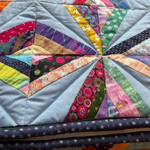 May include: A vibrant, handmade quilt with a starburst pattern. The quilt has a light blue base with radiating strips of patterned fabric in shades of pink, yellow, and purple. The edges are finished with a dark blue border with a patterned design.