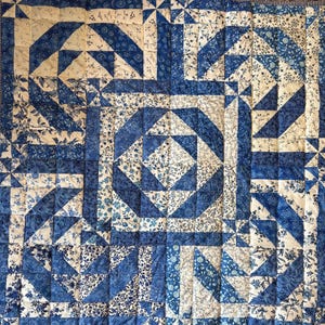 May include: A handmade quilt with a geometric pattern in shades of blue and white. The quilt features various shapes, including squares, triangles, and floral prints. The border has a striped pattern. The quilt is likely made of cotton.