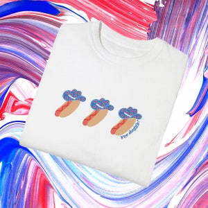 a folded white t-shirt with a graphic of 3 consecutively lined up hot dogs wearing red, white, and blue star-patterned cowboy hats, with the words “Yee doggy” in blue under the third hotdog in the center of the chest area