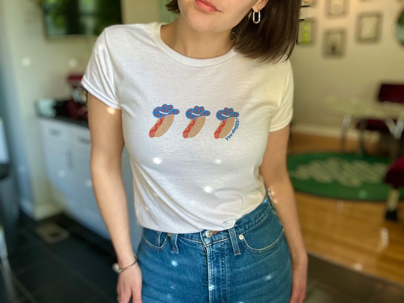 woman wearing a t-shirt with a graphic of 3 consecutively lined up hot dogs wearing red, white, and blue star-patterned cowboy hats, with the words “Yee doggy” in blue