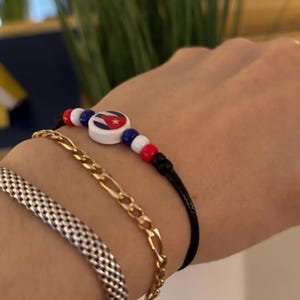 Puede incluir: Una colección de pulseras. Una pulsera de cordón negro con cuentas rojas, blancas y azules y una cuenta central con la bandera cubana. También, una pulsera de cadena dorada y una pulsera de malla plateada.
