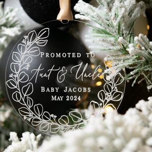 May include: Clear, round Christmas ornament with white text and a floral design. The text reads "Promoted to Aunt & Uncle" with "Baby Jacobs May 2024" below. The ornament hangs from a gold ribbon, set against a backdrop of a Christmas tree.