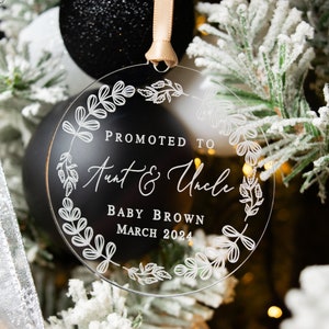 May include: Clear round Christmas ornament with a white floral design and the text "Promoted to Aunt & Uncle Baby Brown March 2024".