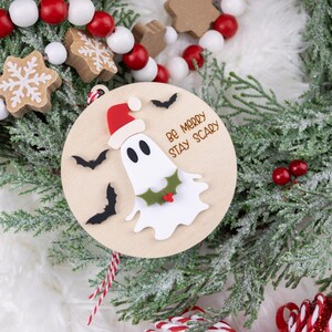 以下が含まれることがあります： サンタ帽をかぶったお化けと「Be Merry Stay Scary」の文字が入った木製のクリスマスオーナメント。オーナメントは黒いコウモリとヒイラギで飾られています。赤と白のビーズガーランドと緑がオーナメントを囲んでいます。