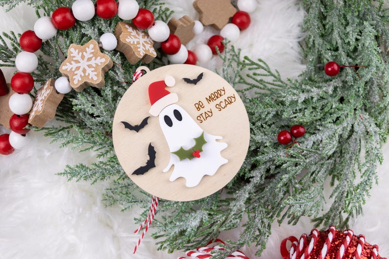 以下が含まれることがあります： サンタ帽をかぶったお化けと「Be Merry Stay Scary」の文字が入った木製のクリスマスオーナメント。オーナメントは黒いコウモリとヒイラギで飾られています。赤と白のビーズガーランドと緑がオーナメントを囲んでいます。