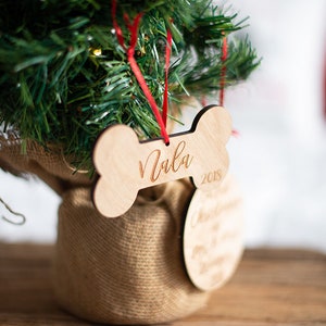 May include: A wooden bone-shaped Christmas ornament with the name "Nala" and the year "2018" engraved on it. The ornament is hanging from a red ribbon on a small artificial Christmas tree.