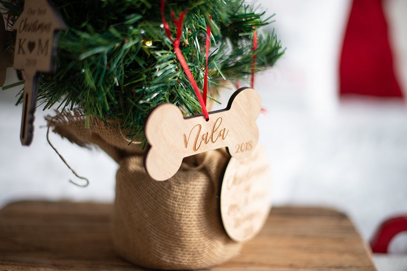 May include: A wooden bone-shaped Christmas ornament with the name "Nala" and the year "2018" engraved on it. The ornament is hanging from a red ribbon on a small artificial Christmas tree.