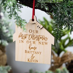 May include: A wooden house-shaped Christmas ornament with the text "Our first Christmas in our new home Brittany & Kevin" and the year "2019" engraved on it.
