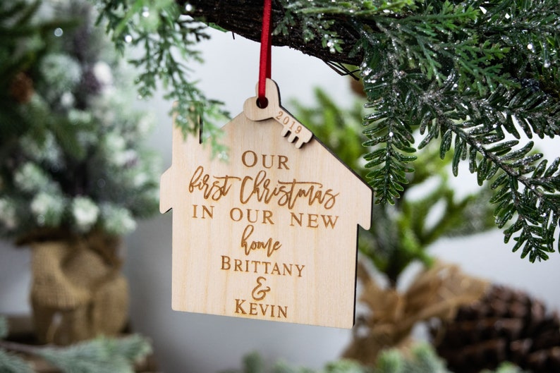 May include: A wooden house-shaped Christmas ornament with the text "Our first Christmas in our new home Brittany & Kevin" and the year "2019" engraved on it.