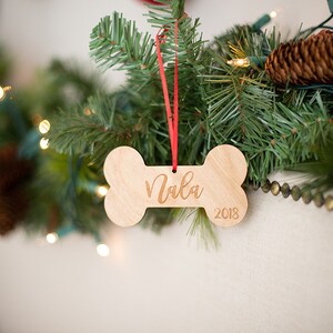 May include: A wooden dog bone ornament with the name "Nala" and the year "2018" engraved on it. The ornament is hanging from a red ribbon on a Christmas tree.