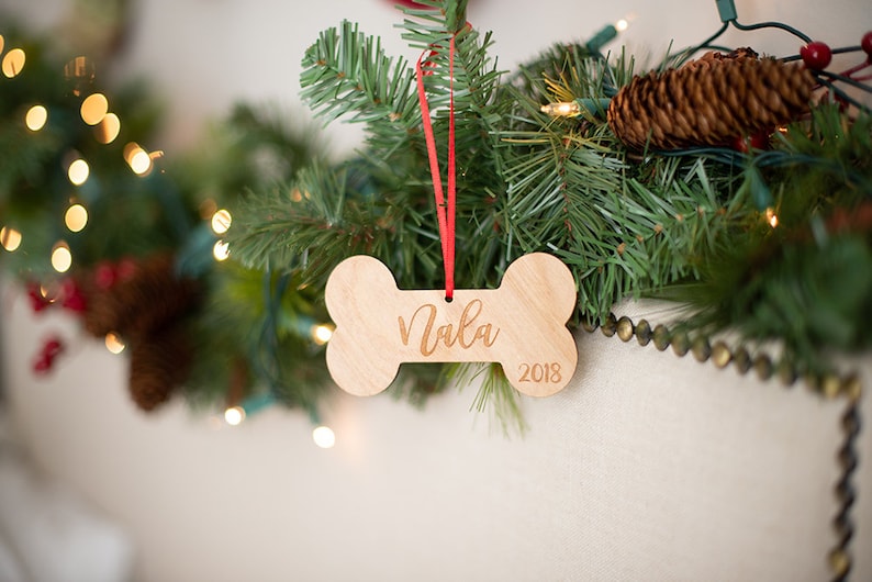 May include: A wooden dog bone ornament with the name "Nala" and the year "2018" engraved on it. The ornament is hanging from a red ribbon on a Christmas tree.
