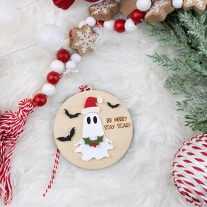 以下が含まれることがあります： サンタ帽をかぶったお化けと「Be Merry Stay Scary」の文字が入った木製のクリスマスオーナメント。赤いと白のビーズ、ジンジャーブレッドクッキー、常緑樹の小枝など、お祝いの装飾が施されています。