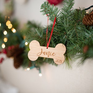 May include: A wooden bone-shaped Christmas ornament with the name "Jamie" and the year "2018" engraved on it. The ornament is hanging from a red ribbon on a green Christmas tree.