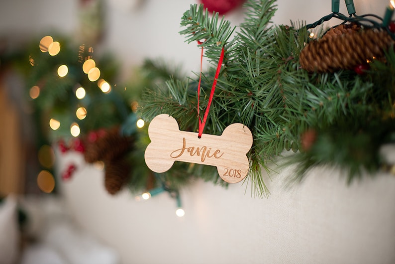 May include: A wooden bone-shaped Christmas ornament with the name "Jamie" and the year "2018" engraved on it. The ornament is hanging from a red ribbon on a green Christmas tree.
