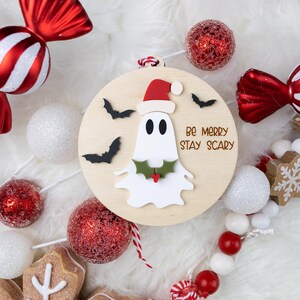 以下が含まれることがあります： サンタ帽をかぶったお化けと「Be Merry Stay Scary」の文字が入った丸い木製オーナメント。オーナメントは、赤と白のキャンディー、オーナメント、ガーランドに囲まれています。