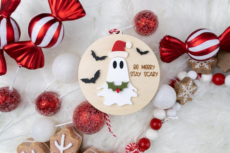 以下が含まれることがあります： サンタ帽をかぶったお化けと「Be Merry Stay Scary」の文字が入った丸い木製オーナメント。オーナメントは、赤と白のキャンディー、オーナメント、ガーランドに囲まれています。