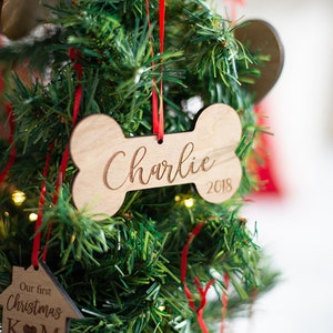 May include: A wooden bone-shaped Christmas ornament with the name "Charlie" and the year "2018" engraved on it. The ornament is hanging on a green artificial Christmas tree.