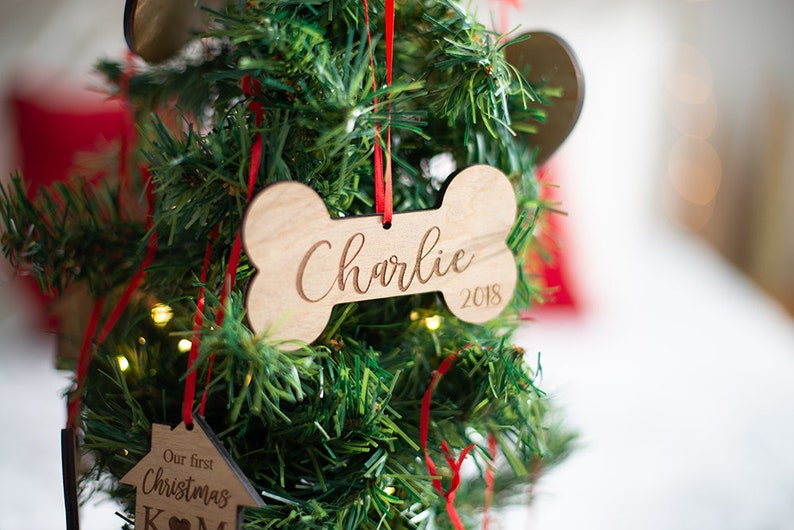 May include: A wooden bone-shaped Christmas ornament with the name "Charlie" and the year "2018" engraved on it. The ornament is hanging on a green artificial Christmas tree.