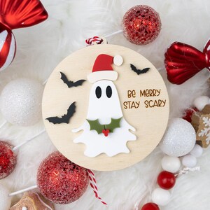 以下が含まれることがあります： サンタ帽をかぶったお化けと「Be Merry Stay Scary」の文字が入った木製のクリスマスオーナメント。キャンディー型のオーナメントやキラキラ光るボールなど、赤と白のホリデーデコレーションに囲まれています。