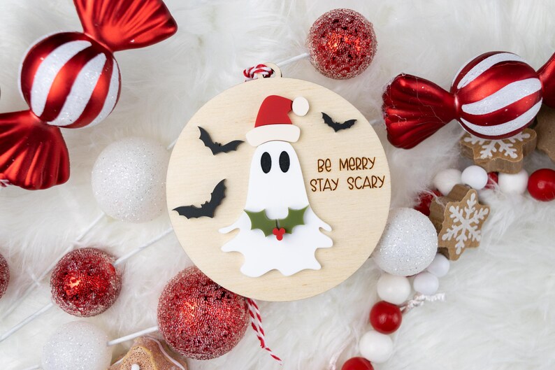 以下が含まれることがあります： サンタ帽をかぶったお化けと「Be Merry Stay Scary」の文字が入った木製のクリスマスオーナメント。キャンディー型のオーナメントやキラキラ光るボールなど、赤と白のホリデーデコレーションに囲まれています。