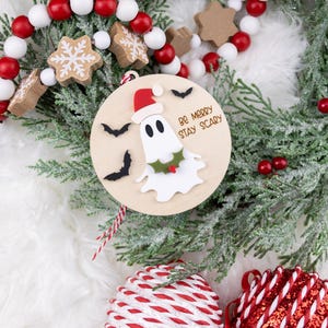以下が含まれることがあります： サンタ帽をかぶったお化けと「Be Merry Stay Scary」の文字が入った木製のクリスマスオーナメント。オーナメントは黒いコウモリとヒイラギで飾られています。お祝いの休日の装飾。