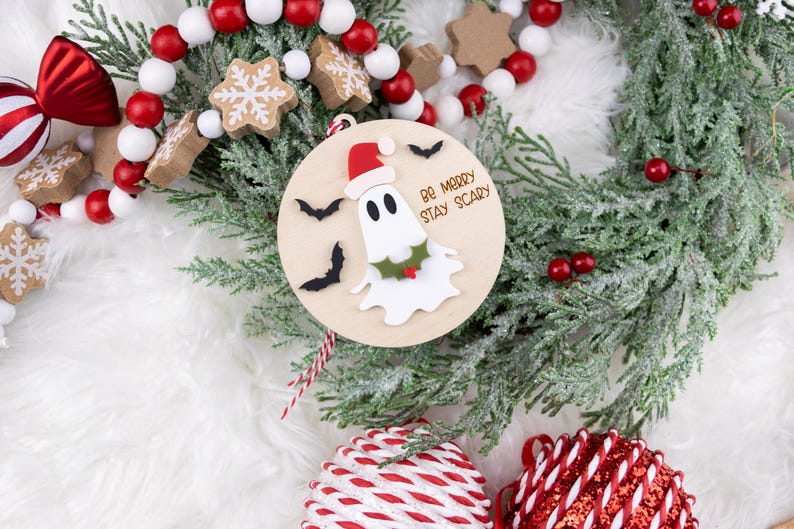 以下が含まれることがあります： サンタ帽をかぶったお化けと「Be Merry Stay Scary」の文字が入った木製のクリスマスオーナメント。オーナメントは黒いコウモリとヒイラギで飾られています。お祝いの休日の装飾。